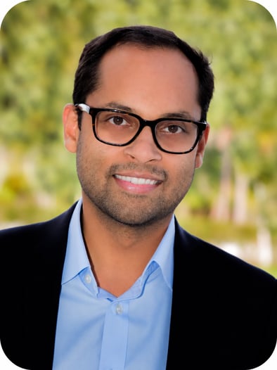 Sanjeev Joseph smiling in a professional headshot, wearing glasses, a dark blazer, and a light blue shirt against a softly blurred outdoor background.