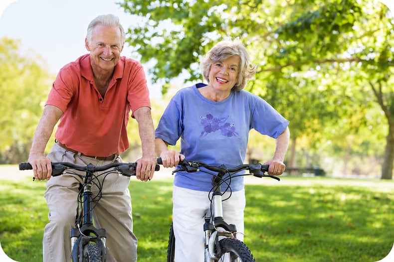An elderly man and woman out in a green park with a tree, on their bikes, smiling because physical therapy supports their healthy lifestyle. 