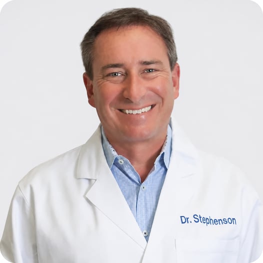 FYZICAL's Dr. Carl Stephenson, smiling in a white medical coat over a blue collared shirt.