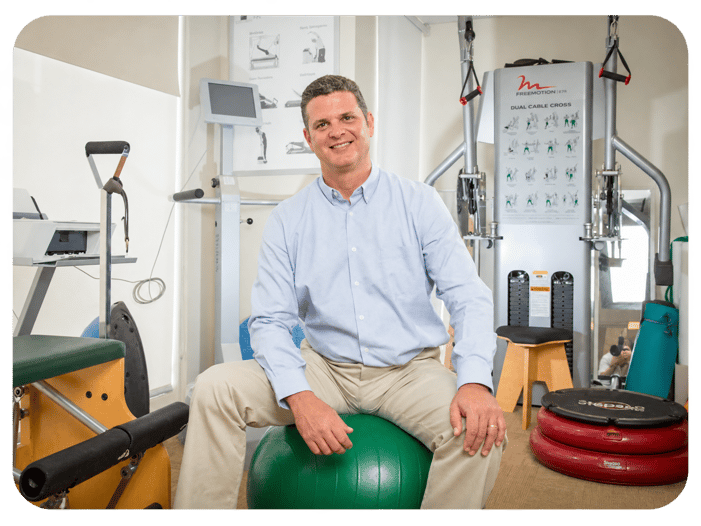 Alex Matz sitting on an exercise ball inside a FYZICAL physical therapy clinic surrounded by rehabilitation equipment.