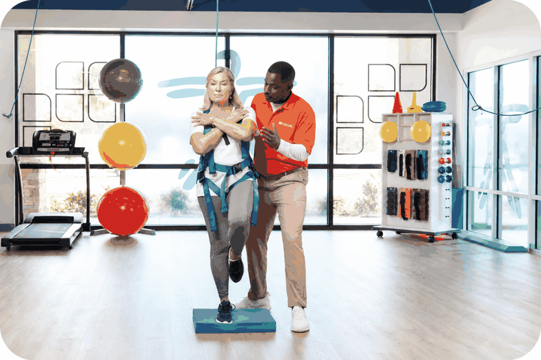 A physical therapist assisting a patient with balance training on a foam pad in a FYZICAL clinic.