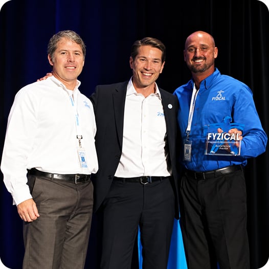 Three FYZICAL team members, including Chris Brunelle and Mike Shiver, posing together at an event, with one holding a FYZICAL Excellence in Practice award.