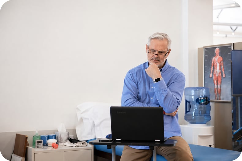 An elderly man in a blue long sleeve shirt looking intently at a laptop while getting information from telehealth about physical therapy. 