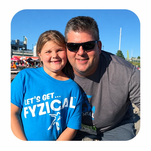 Eric Bjorkman smiling with a young girl wearing a blue FYZICAL T-shirt at an outdoor community event.