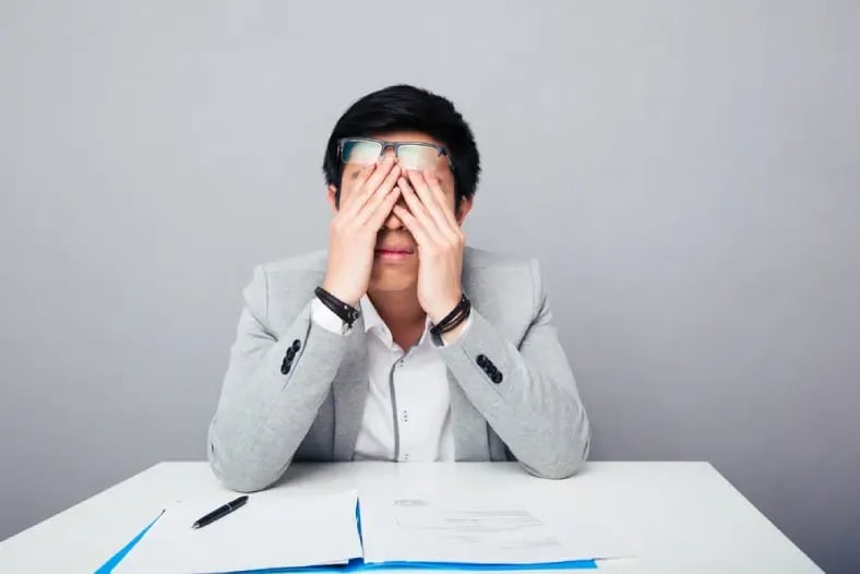 Young asian businessman sitting at the table and rubbing his eyes over gray background