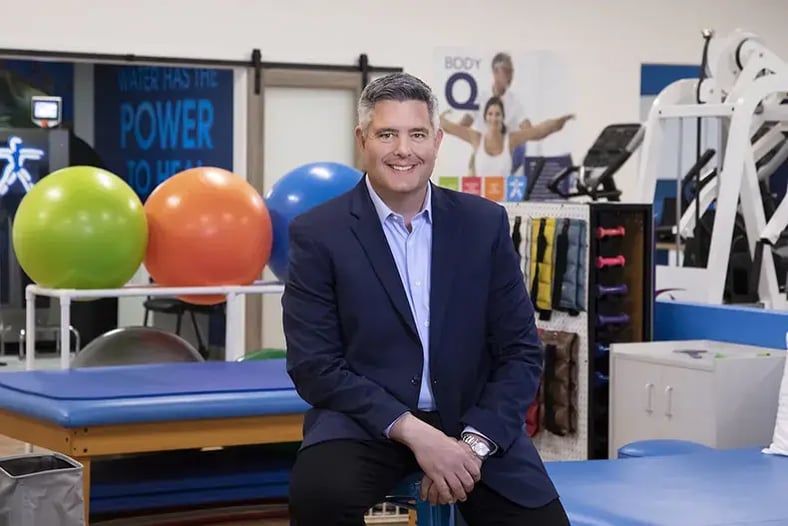 Franchisee smiling and sitting in front of therapy equipment